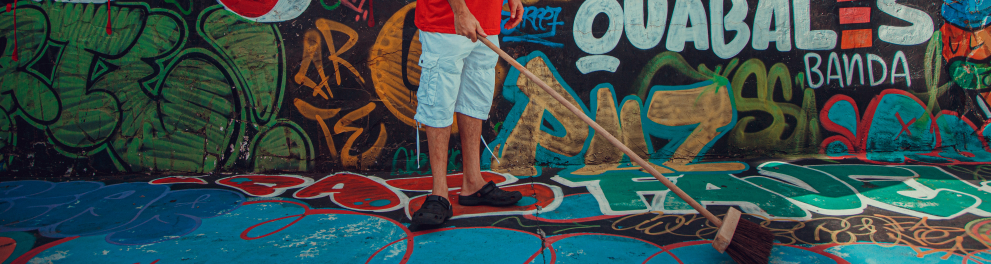 Um homem negro, de cabelo dread comprido, varre o chão. Ao fundo, uma parede pintada com grafite.