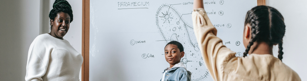 À esquerda, uma mulher negra sorri ao ver as duas crianças interagindo com ela em uma sala de aula. Ela é a professora. O menino (ao centro) e a menina (à direita), os estudantes.