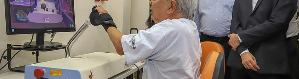 Um idoso branco está com um acessório robótico no braço esquerdo enquanto é observado por outros homens trajando paletó ao lado