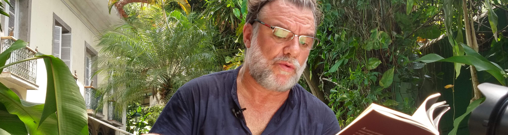 Um homem, com cabelo e barba grisalhos, segura um livro. Ao fundo, plantas.