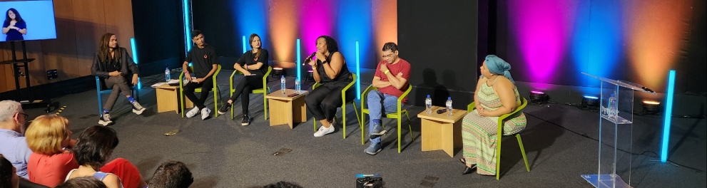 Pessoas estão sentadas em cadeiras sobre um palco, em um auditório.