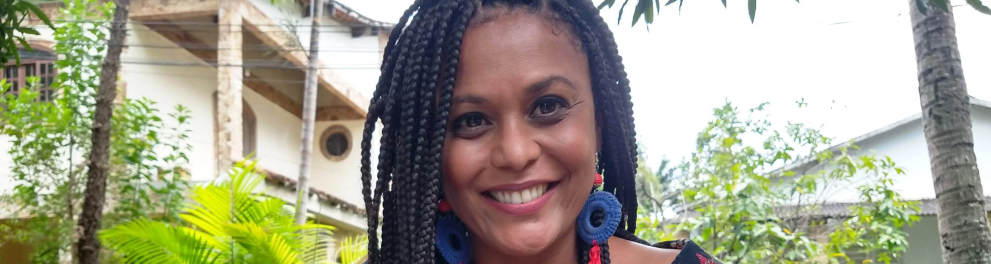 Mulher negra sorri. Ao fundo, o céu e algumas plantas.