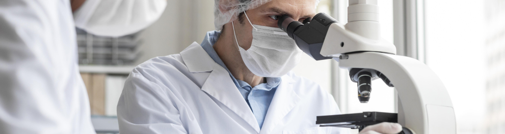 Cientistas de jaleco, máscara de proteção contra covid, luvas e toucas protetoras estão em um laboratório. Um deles está olhando algo em um microscópio