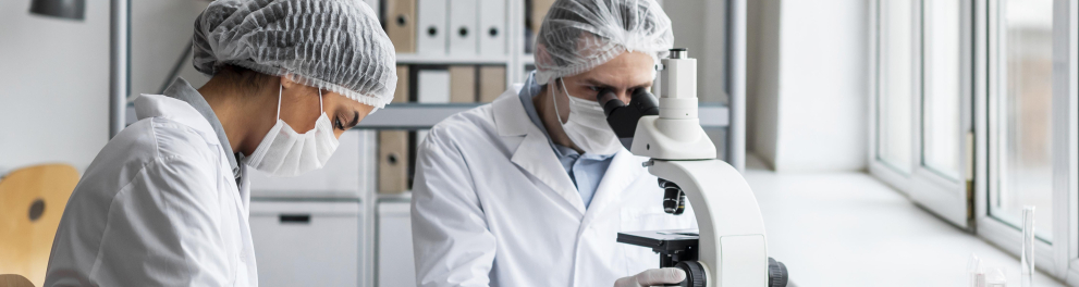 Cientistas de jaleco, máscara de proteção contra covid, luvas e toucas protetoras estão em um laboratório. Um deles está olhando algo em um microscópio