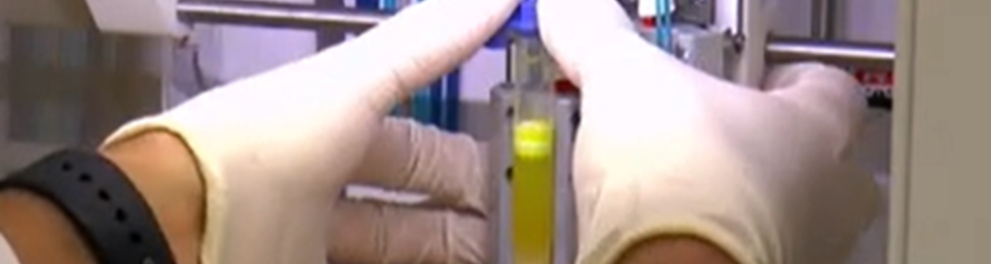 Mãos humanos com luvas brancas de borracha estão tocando um aparelho eletrônico, que contém um recipiente de vidro. Trata-se de um ambiente de laboratório