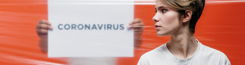 Uma mulher branca de perfil está à frente de um homem segurando um cartaz com a palavra CORONAVIRUS escrita. Há um plástico filme entre as duas pessoas