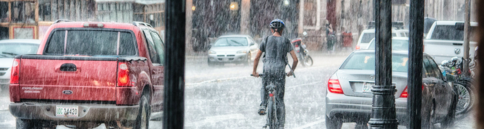 Imagem de uma enchente caindo sobre uma zona urbana, com carros e um ciclista em meio à forte chuva
