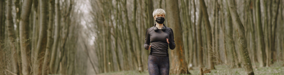 Mulher de cabelos curtos e loiros está de máscara de proteção à covid-19, roupa de malhação e correndo em uma área ao ar livre arborizada