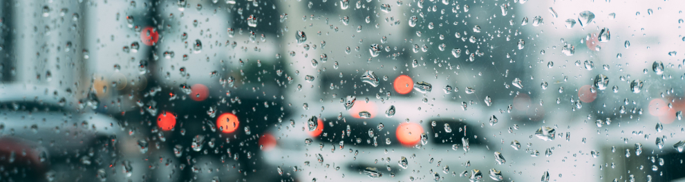 Imagem embaçada feita por trás de um vidro molhado de chuva. Podemos observar uma rua com vários automóveis andando durante chuva intensa