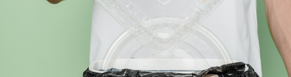 Uma pessoa usando luva descartável está jogando um recipiente de plástico em uma lixeira com a palavra "plastic" escrita