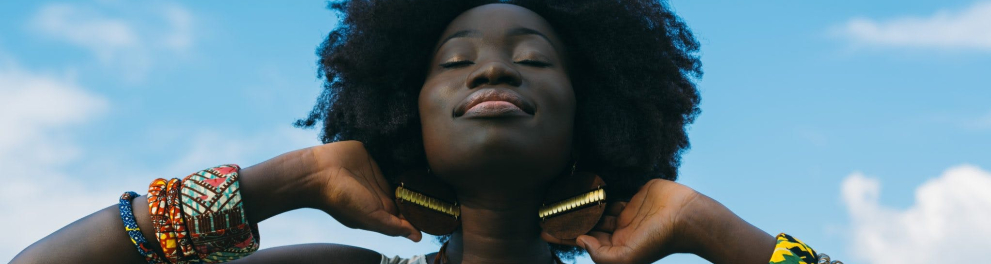 Uma mulher negra está com as mãos atrás da cabeça, como se estivesse se espreguiçando, dando um leve sorriso, com os olhos fechados. Ao fundo, o céu azul.