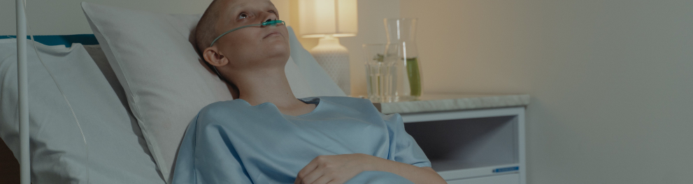 Uma mulher jovem está deitada em uma cama de hospital com tubos de oxigênio. A imagem é em preto e branco e a pessoa está careca, o que nos passa a ideia de ser uma paciente com câncer