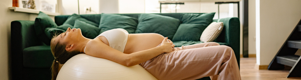 Uma mulher branca e grávida está deitada sobre uma bola de yoga, com as duas mãos na barriga. Ela parece estar fazendo algum tipo de yoga para gestantes