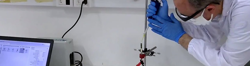 Cientista está de jaleco branco e luvas azuis de borracha mexendo em um sensor cilíndrico de metal