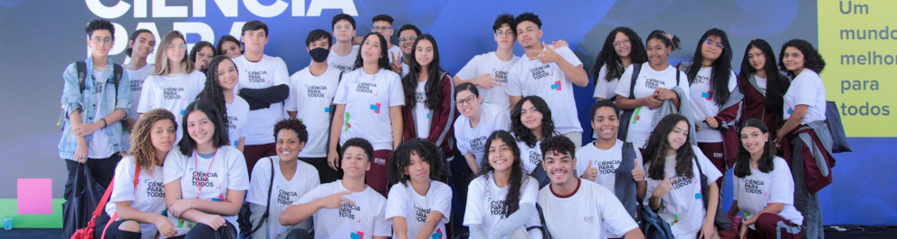 Várias crianças uniformizadas posando em frente a um banner azul do "Ciência para todos"