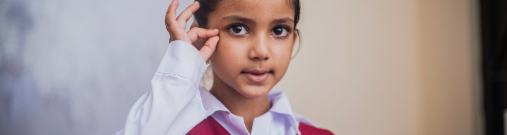 Uma menininha de traços indianos está fazendo um sinal com a mão direita como se estivesse se comunicando pela linguagem de libras