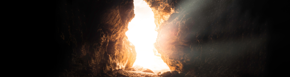Imagem de uma abertura entre rochas, que é a saída de uma caverna escura...essa abertura está visível graças á luz solar