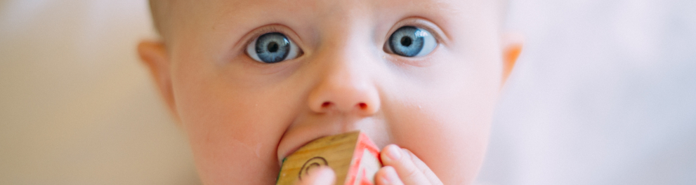 Close em um bebê branco de olhos azuis e cabelos loiros, que está segurando um objeto com as duas mãos