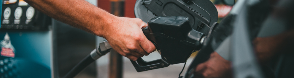 Imagem em zoom da mão de uma pessoa branca colocando uma mangueira de gasolina no tanque de um automóvel estacionado em um posto de gasolina