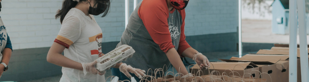 Duas mulheres estão de máscaras e luvas de proteção à covid-19 mexendo em sacolas de papelão com alimentos como ovos, por exemplo