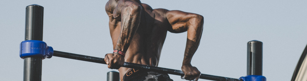 Um jovem homem negro bem musculoso está sem camisa fazendo o exercício chamado "paralela" em traves de ferro ao ar livre