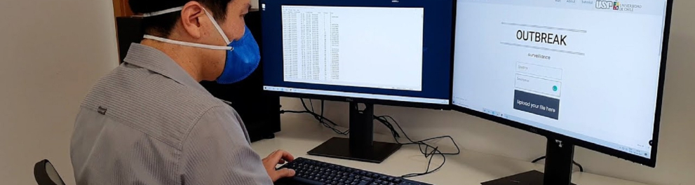 Um homem de máscara de proteção à Covid 19 está sentado trabalhando diante de um computador com dois monitores