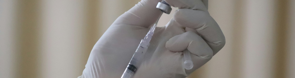 Uma injeção sendo manuseada por mãos trajadas de luvas brancas