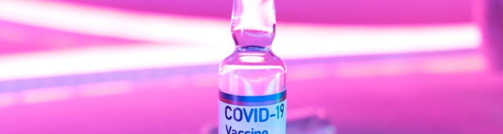 Um vidrinho contendo um líquido transparente está em cima de uma mesa e há uma rótulo envolvendo o frasco, com os dizeres:  "covid-19 vaccine"