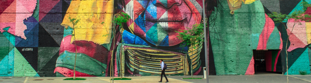 Um homem caminha na calçada, em frente a uma grande parede de um prédio em que um homem indígena está pintado, em diversas cores.