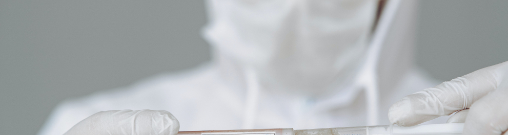 Um cientista branco está de óculos e máscara de proteção, além de luvas e jaleco branco, mexendo em dois recipientes de vidro com um líquido, como se estivesse passando o líquido de um recipiente para outro