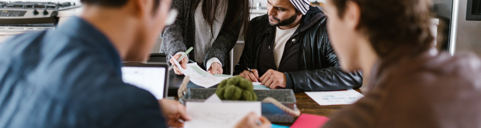Adultos jovens estão sentados a uma mesa discutindo sobre aspectos que veem em folhas de papel.