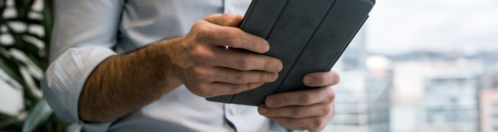 Imagem de um homem segurando um tablet com as duas mãos. Ele está de roupa social e a cabeça não aparece, é uma imagem apenas do tronco focando no objeto que segura
