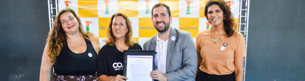 Quatro pessoas (três mulheres e um homem) estão de pé, sorrindo, segurando o documento assinado para firmar a parceria da co.liga com a prefeitura de São Vicente.