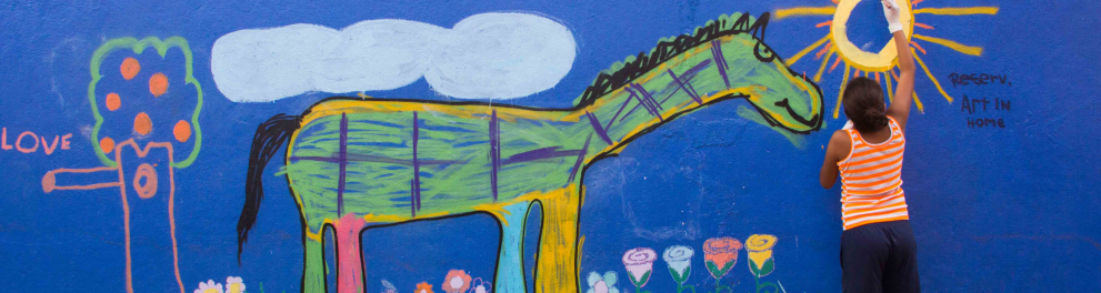 Uma menina desenha um sol amarelo em uma parede azul. Na parede já estão desenhados um cavalo, um gramado com flores e uma árvore.