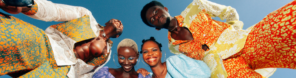 Quatro pessoas negras (dois homens, nas pontas, 2 mulheres, no meio) estão de pé, vestidas com roupas coloridas. Todas sorriem. A fotografia foi tirada de baixo pra cima, com elas e eles olhando pra baixo (pra quem os fotografou).