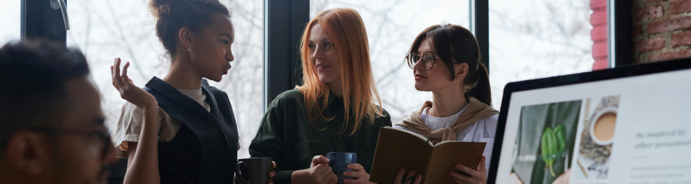 Três meninas jovens estão de pé, conversando, enquanto um rapaz está sentado, utilizando um computador.
