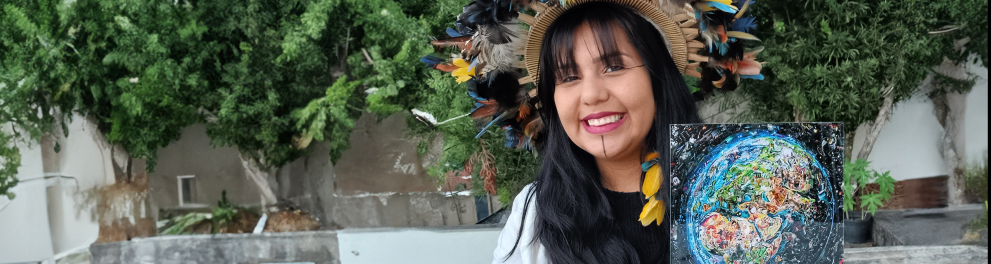 Uma mulher indígena sorri com um buquê nas mãos. Ao fundo, folhas de uma árvore.