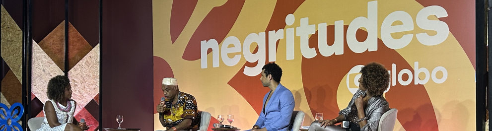 Quatro pessoas negras estão sentadas em poltronas sobre um palco, diante de uma plateia. Ao fundo, sobre uma parede em tons de marrom-claro, o nome do evento: Negritudes Globo.