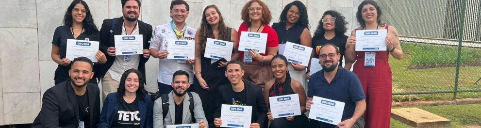 Pessoas de pé e agachadas (à sua frente) seguram diplomas enquanto sorriem.