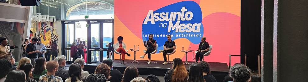 Quatro pessoas estão sentadas em poltronas sobre um palco. Ao fundo, colorido, o nome do programa que abordavam ali: Assunto na Mesa.