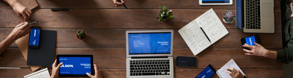 Diversos computadores dispostos sobre uma mesa com algumas pessoas sentadas ao redor dela.