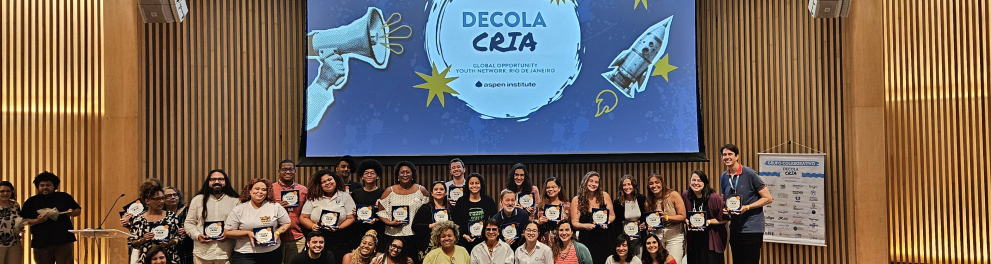 Diversas pessoas (de pé e agachadas, em duas fileiras) sobre um palco. Ao fundo, projetado em tom azulado, o texto "Decola Cria".
