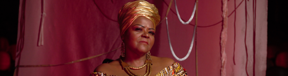 Uma mulher está vestida com uma roupa e um turbante dourados. Ao fundo, um pano rosa.