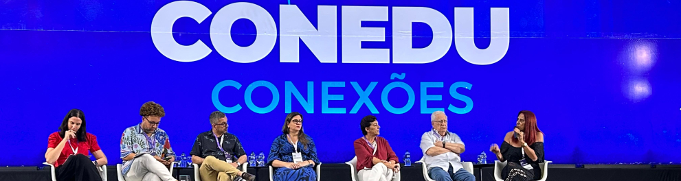 Sete pessoas sentadas em cadeiras sobre um palco. Ao fundo, uma projeção azul com o texto "Conedu conexões" em branco.