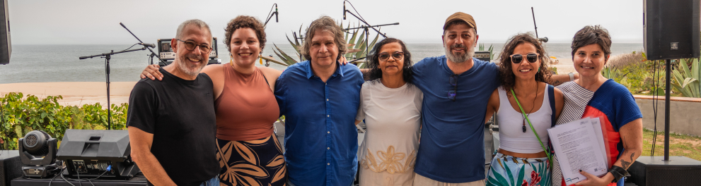Pessoas de pé, sorrindo. Ao fundo, aparelhos de som e o mar.