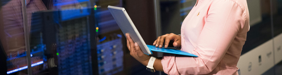 Uma mulher está de pé, com um laptop em suas mãos. Ao fundo, aparelhos tecnológicos.