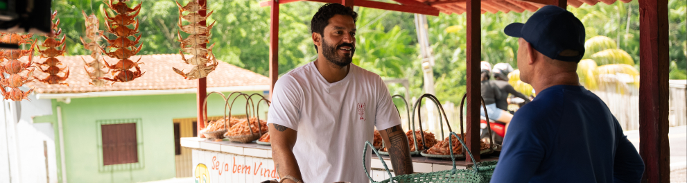 Dois homens estão sorrindo, um para o outro, diante de uma mesa comprida, repleta de alimentos, entre eles.