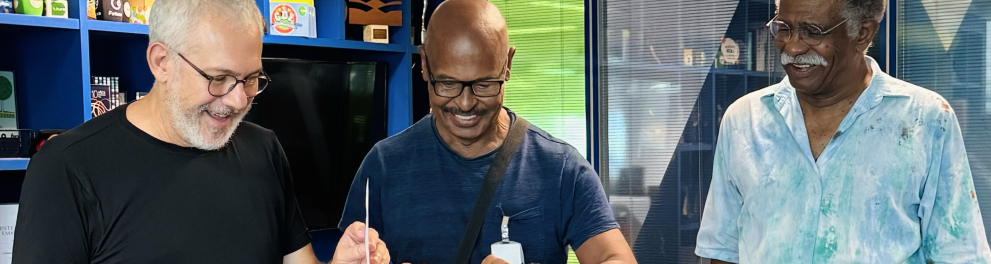 Três homes estão sorrindo, de pé, assinando um termo sobre um púlpito.