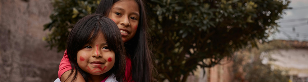 Duas meninas estão abraçadas, olhando para quem as fotografou. Ambas sorriem. Ao fundo, uma árvore e, mais distante, uma casa.
