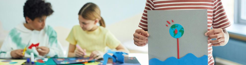 A imagem mostra três estudantes. Um deles está em pé, sorrindo para a foto e segurando um desenho nas mãos. 